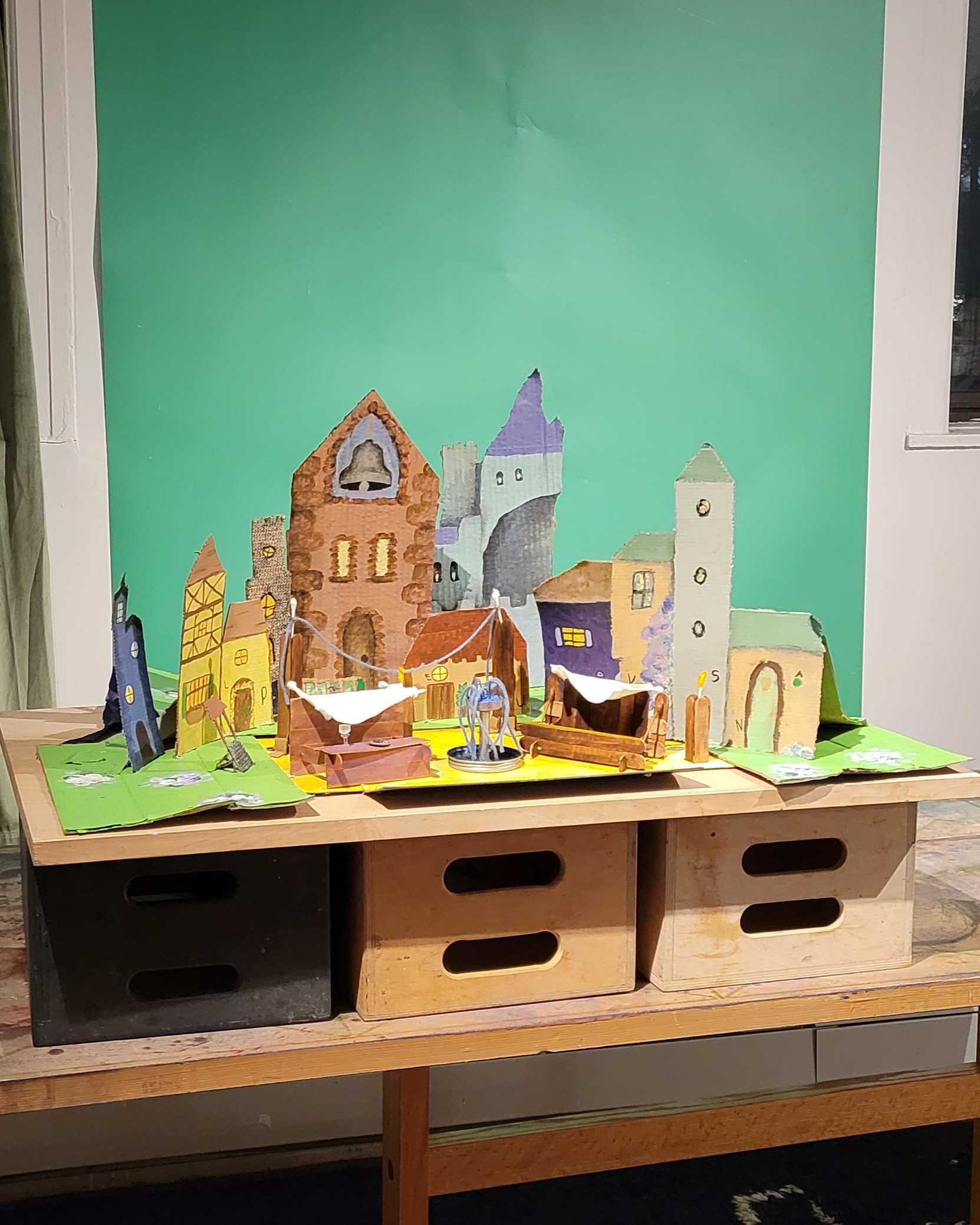 A miniature village of painted cardboard buildings around a market square with stalls made of popsicle sticks.