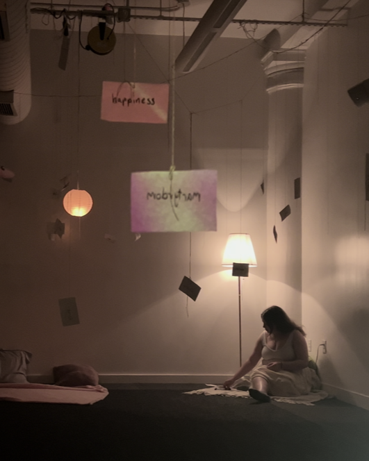 A room with white walls and low, warm lighting, with index cards dangling from the ceiling on strings. A woman sits in the corner under a lamp.