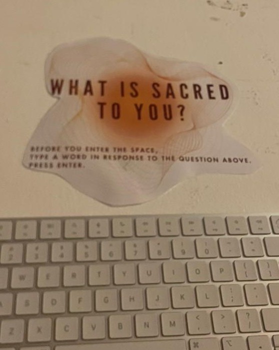 A keyboard with a paper sign above it that reads, 'What is sacred to you? Before you enter the space, type a word in response to the question above. Press enter'.